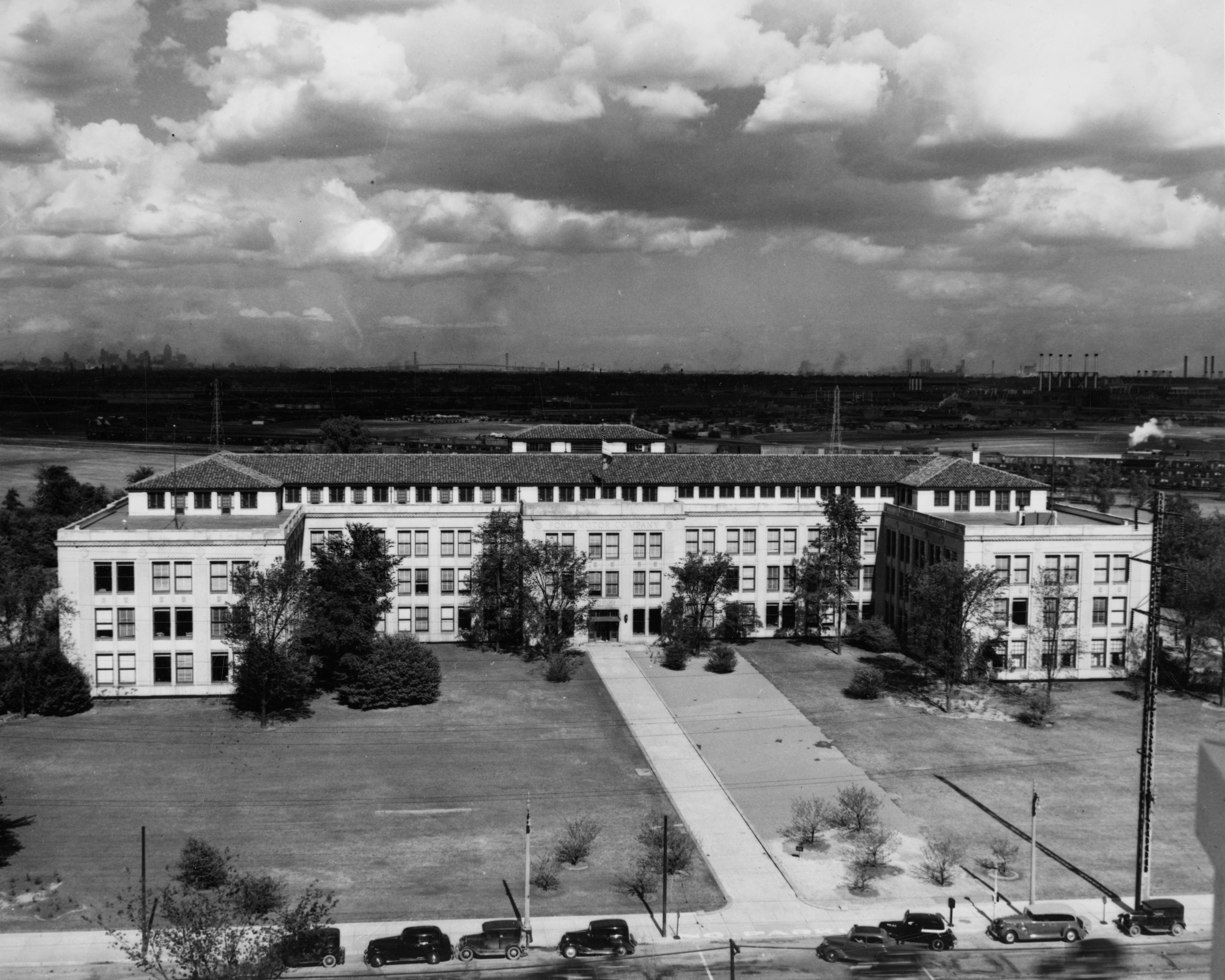 A black and white photo of a large, light-colored building that wraps around a lawn.
